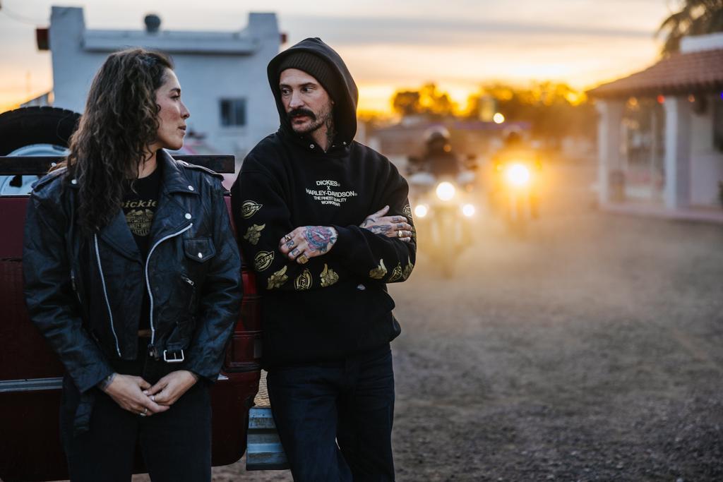 Two people wearing Harley-Davidson gear standing next to a truck with a couple of oncoming motorcycles in the background.